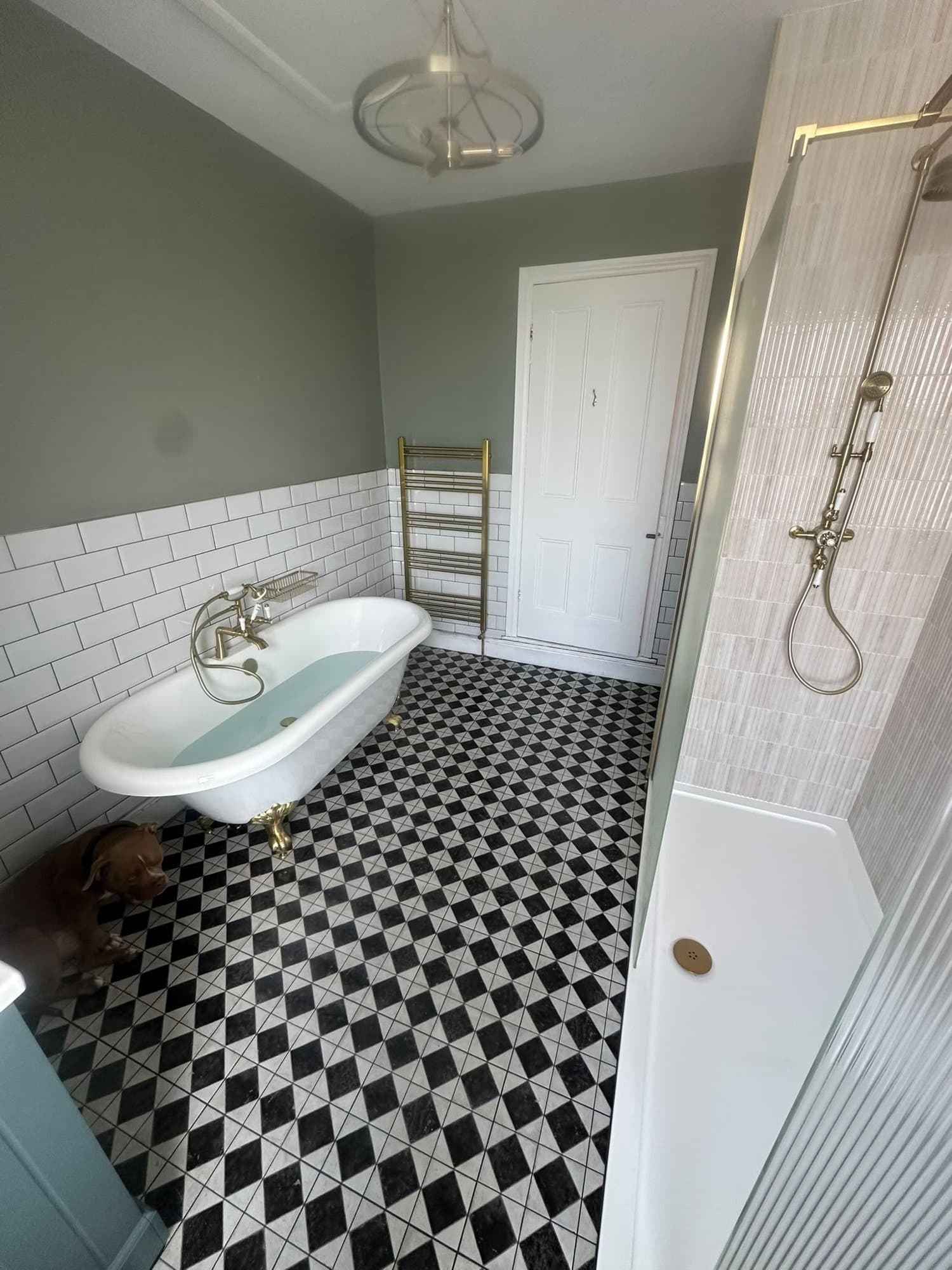 A sage green bathroom with a roll-top bath, brass fittings and a chequered floor
