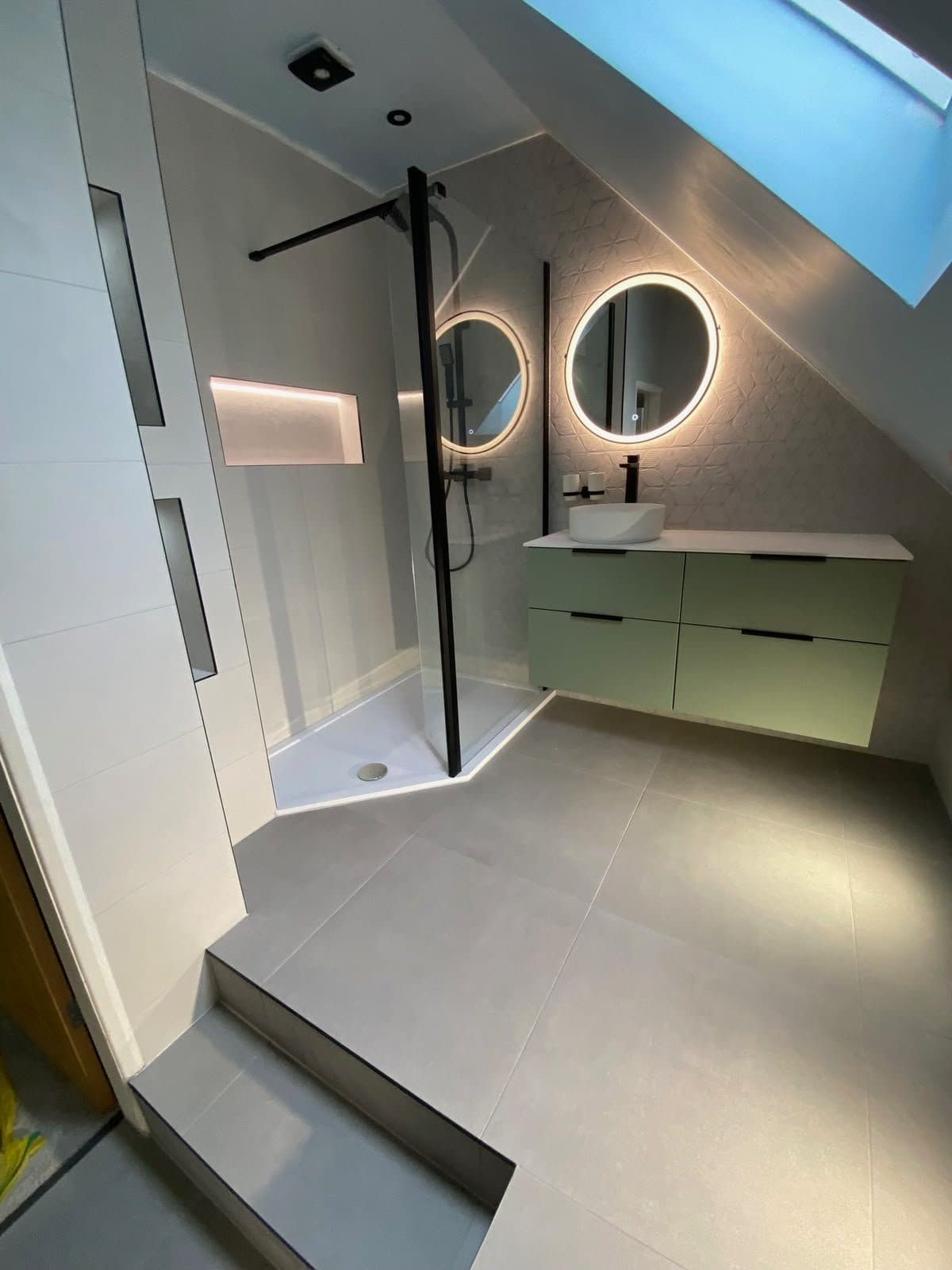 A loft en-suite with dark tiles, a walk-in shower and two round backlit mirrors