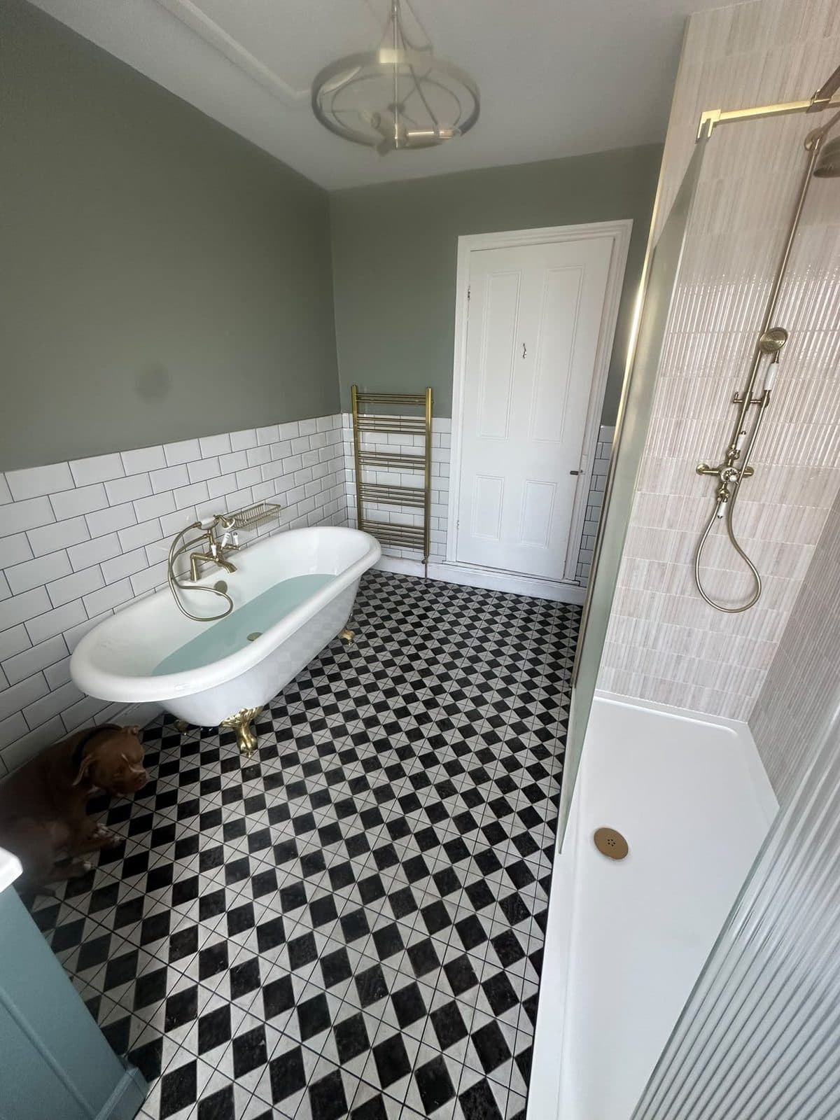 A sage green bathroom with a roll-top bath, brass fittings and a chequered floor