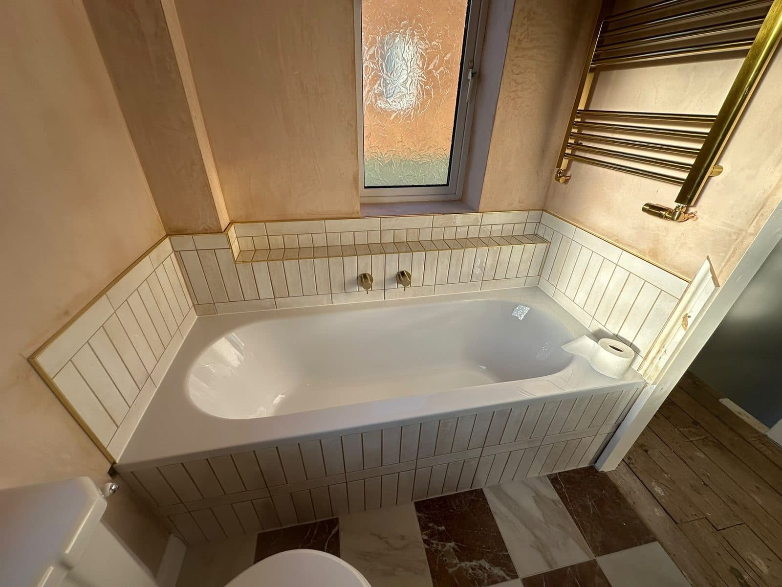 A bath set into a tiled alcove with wood-effect floor tiles and a brass towel radiator
