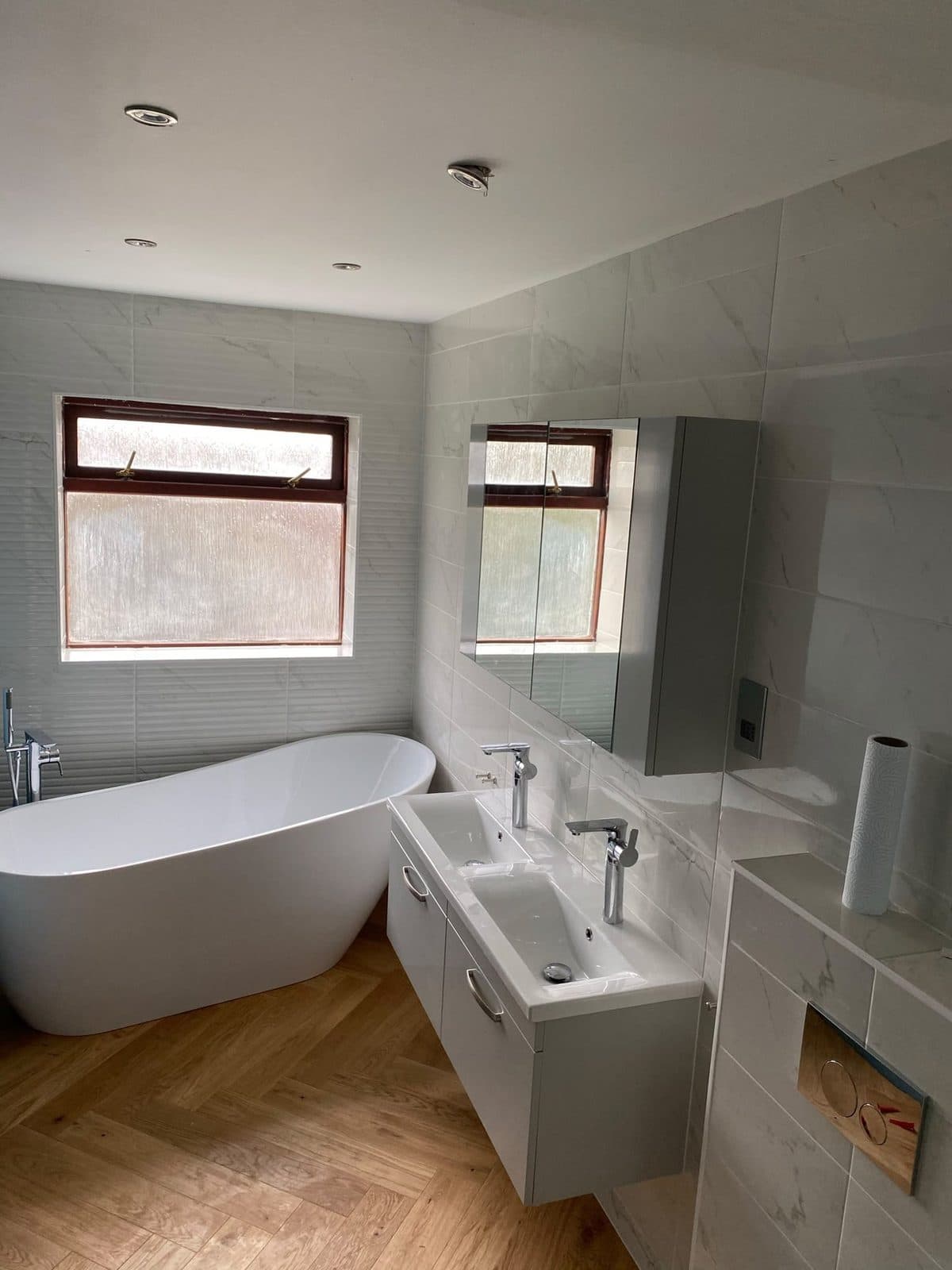 A freestanding bath, wall-hung vanity and tall cabinet on a herringbone wood floor